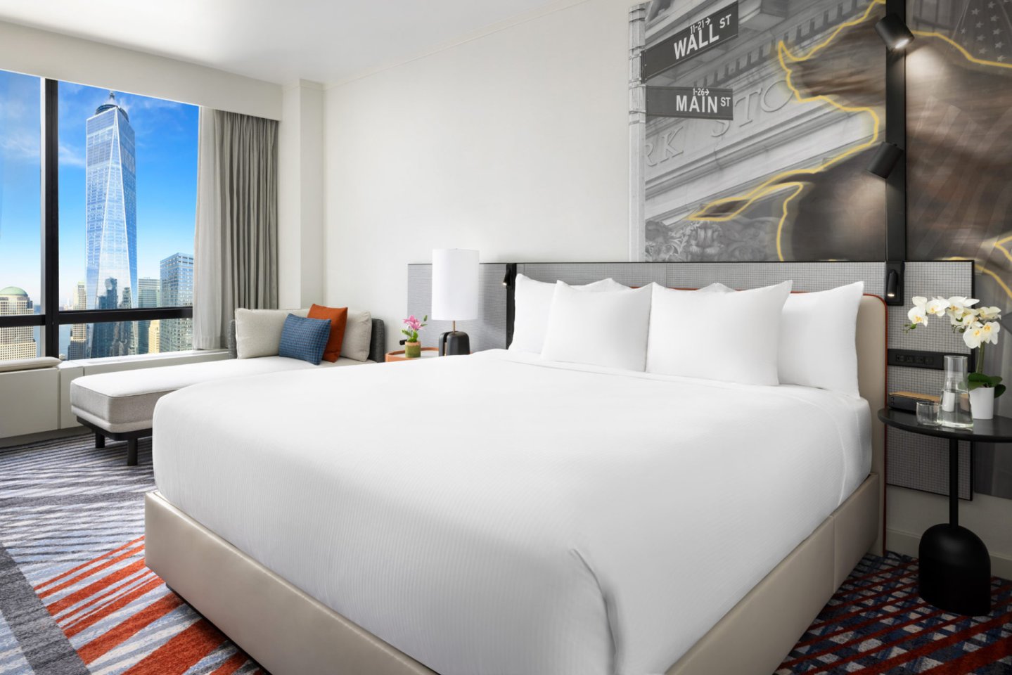 Modern hotel room with a large bed, white linens, a chaise by the window, city views including a tall skyscraper, abstract wall art, and flowers on a nightstand. Natural light brightens the space.