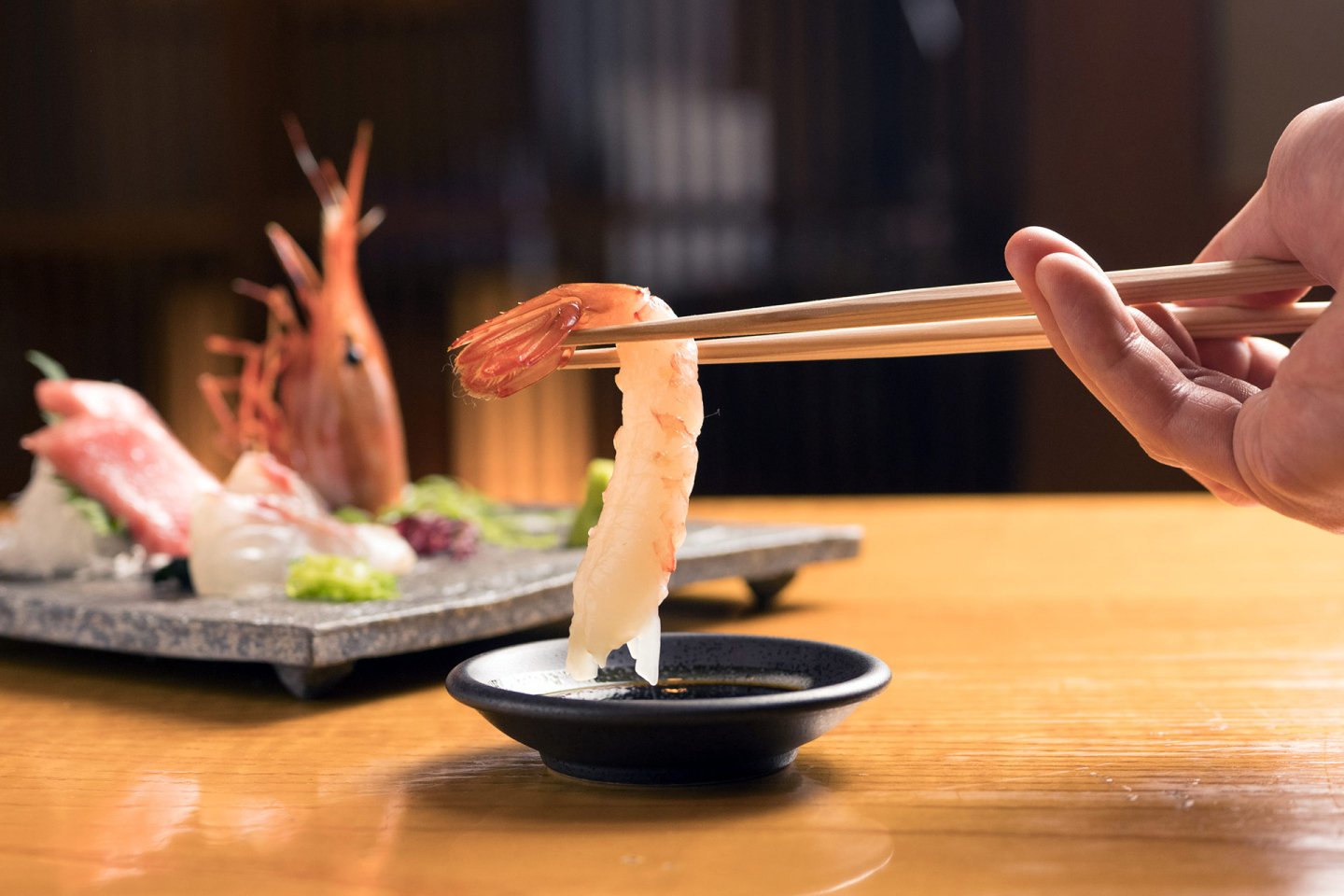 A hand uses chopsticks to dip a piece of shrimp sushi into a small dish of soy sauce, with a plate of assorted sashimi and sushi in the blurred background on a wooden table.