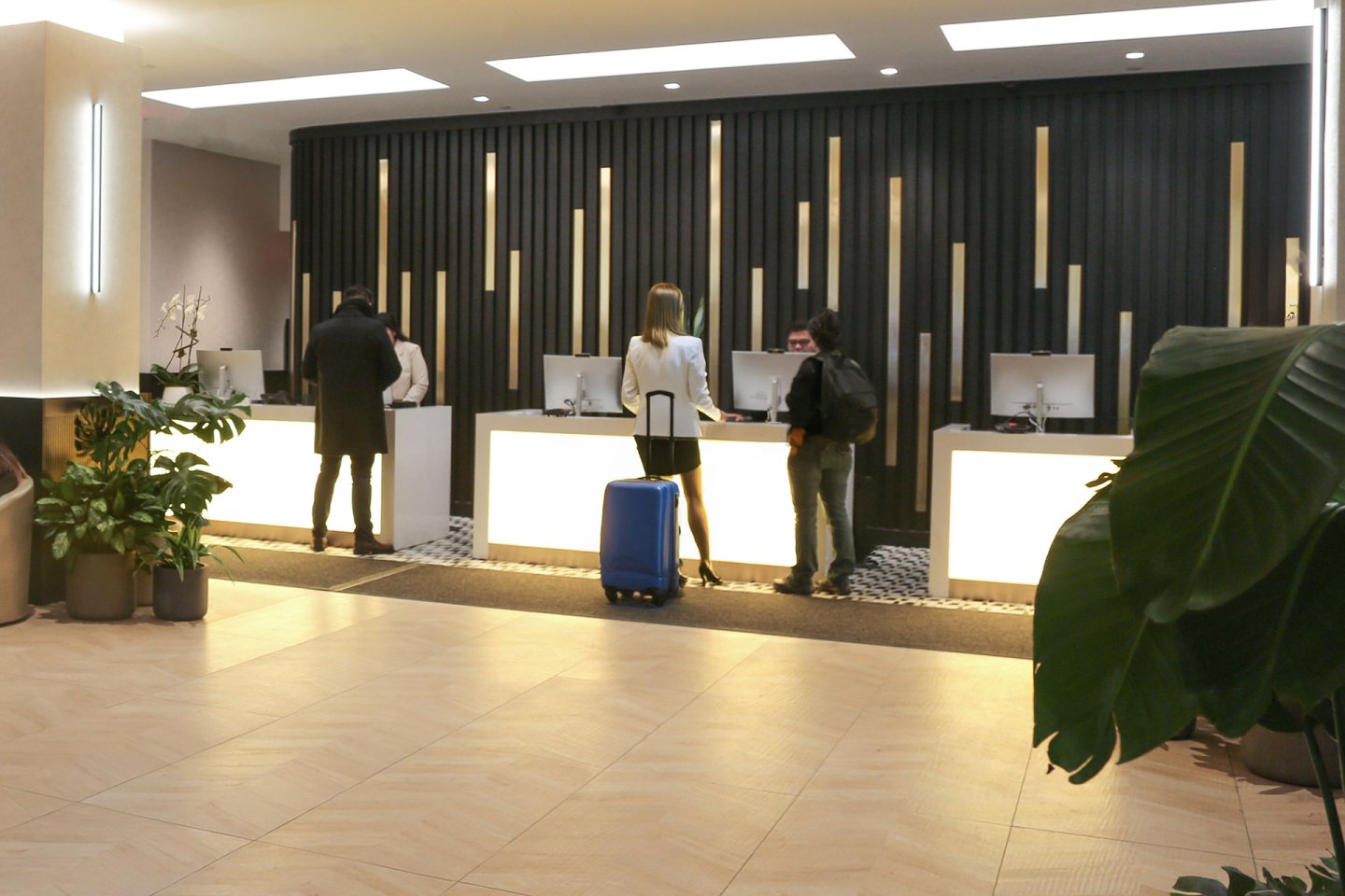 Three people stand at a hotel reception desk with suitcases, speaking to staff. The modern lobby features plants, beige floors, and illuminated counters against a black and gold accent wall.