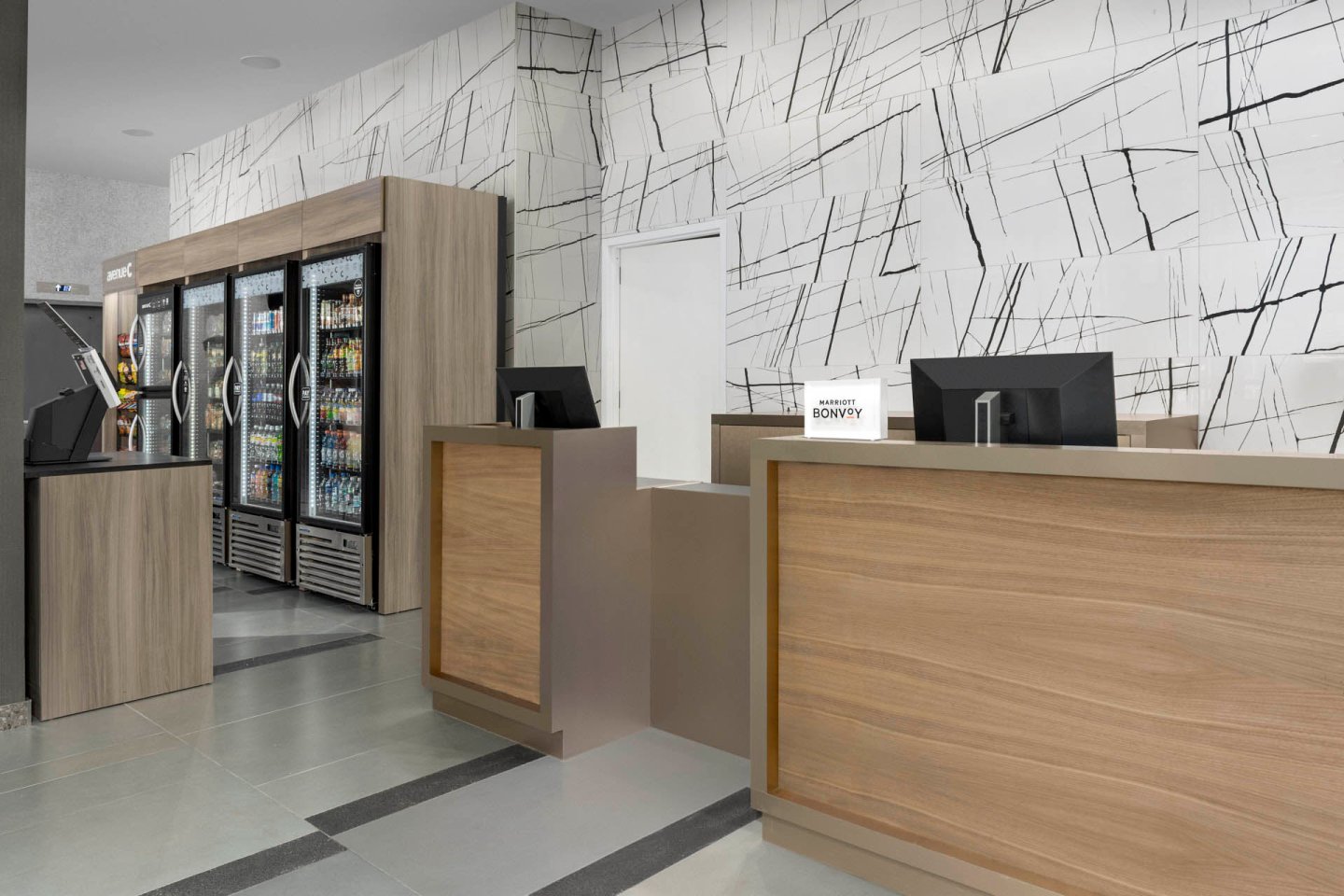 A modern hotel reception area with a wooden desk, computer monitors, and a small sign that reads "Bonvoy." On the left, there is a convenience section with refrigerated drinks and snacks in glass-door coolers