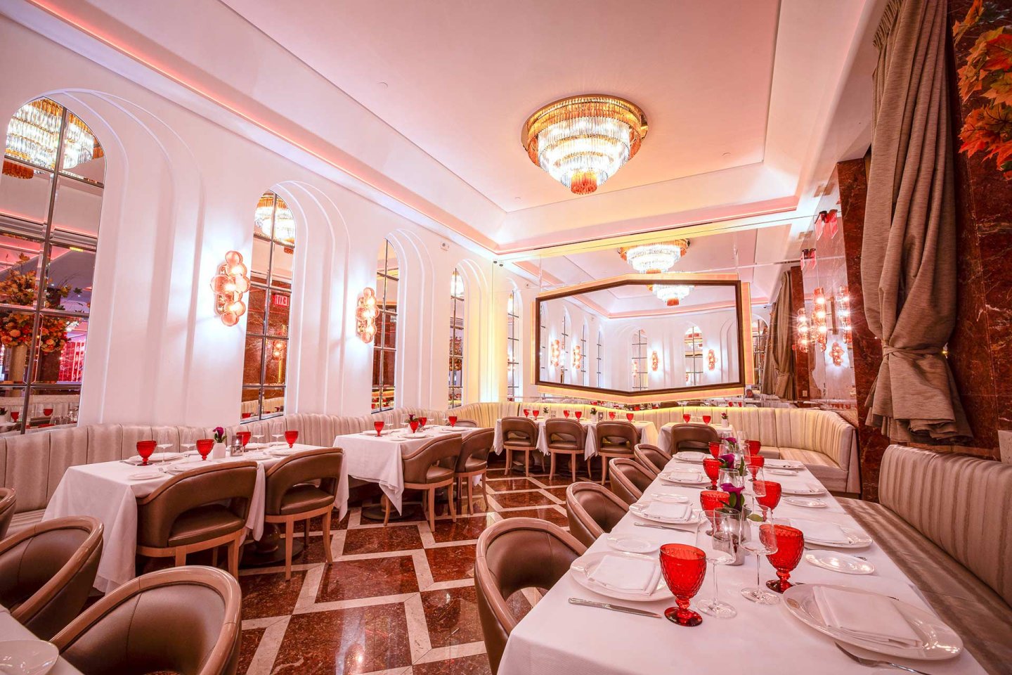 Elegant restaurant interior with chandeliers, white walls, and large mirrors. Tables are set with white cloths, red glasses, plates, and silverware. Tan cushioned chairs and booth seating line the room.