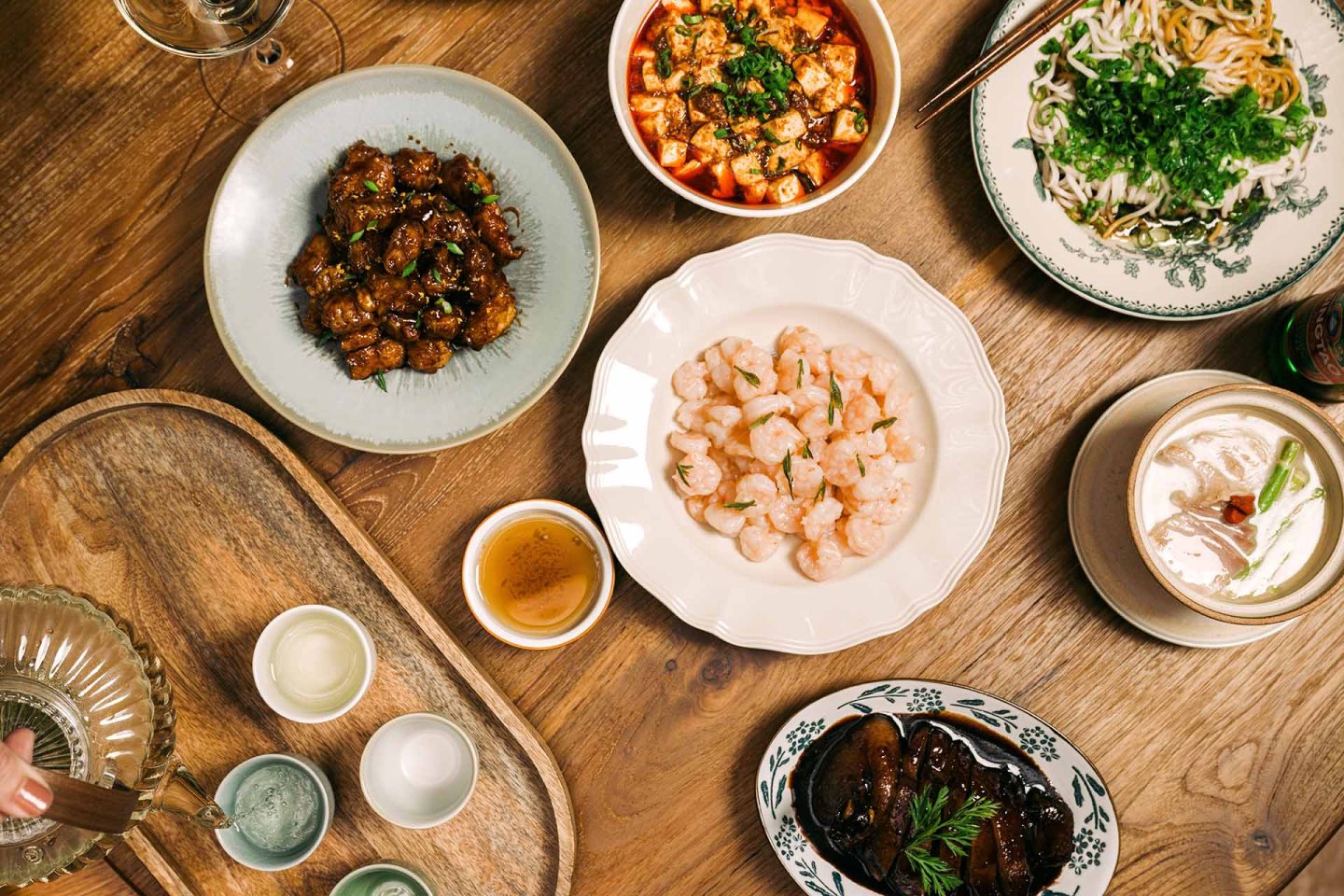 A wooden table set with various Chinese dishes, including shrimp, braised meat, tofu, eggplant, noodle soup, small bowls of sauces, and glasses of clear liquid, with a hand pouring a drink on a tray.