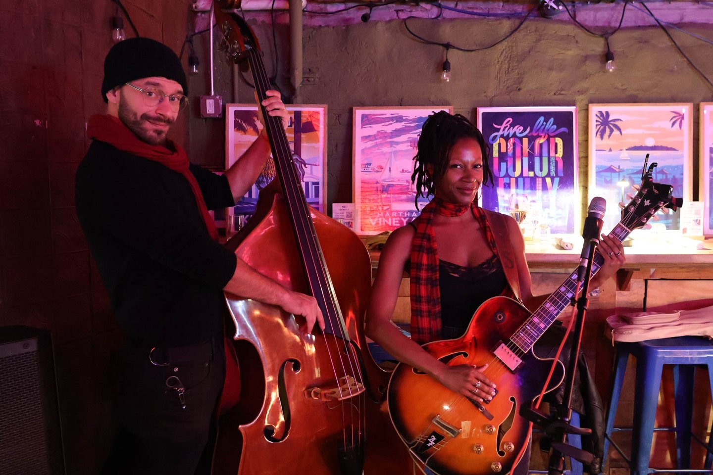 Two musicians stand indoors; one plays an upright bass, wearing a beanie and glasses, and the other holds an electric guitar, wearing a red plaid scarf. Colorful posters and bar stools line the background.