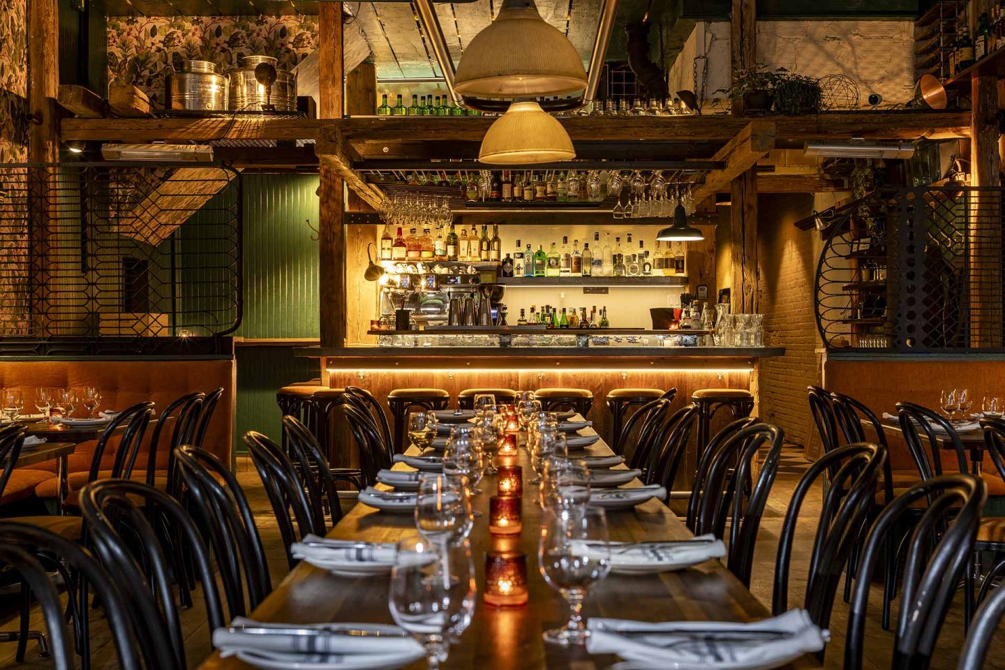 A cozy, dimly lit restaurant interior with long wooden tables set with glasses, plates, and napkins. Candlelight glows along the table. In the background, a well-stocked bar is framed by rustic wooden beams and warm lighting.