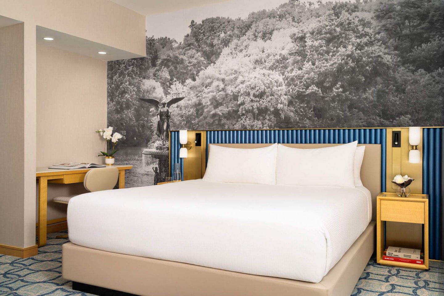 A modern hotel room with a large bed, white bedding, two nightstands with flowers, a desk and chair, and a wall mural featuring a black-and-white image of trees and a statue.