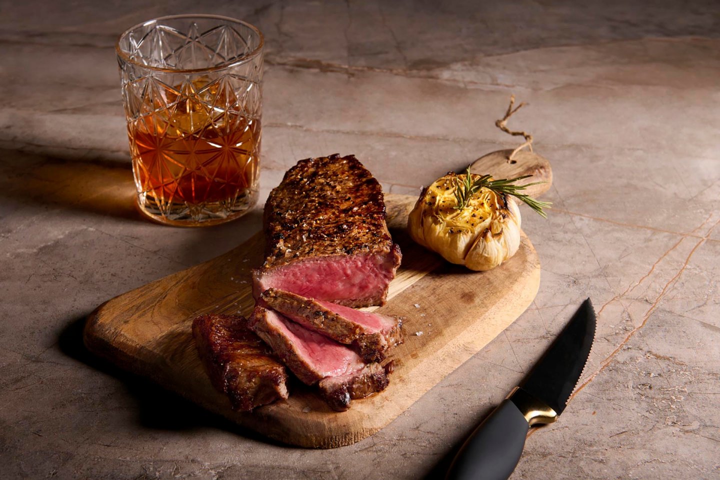 A glass of amber whiskey sits beside a wooden board with sliced medium-rare steak and roasted garlic garnished with rosemary. A knife rests next to the board on a marble surface.