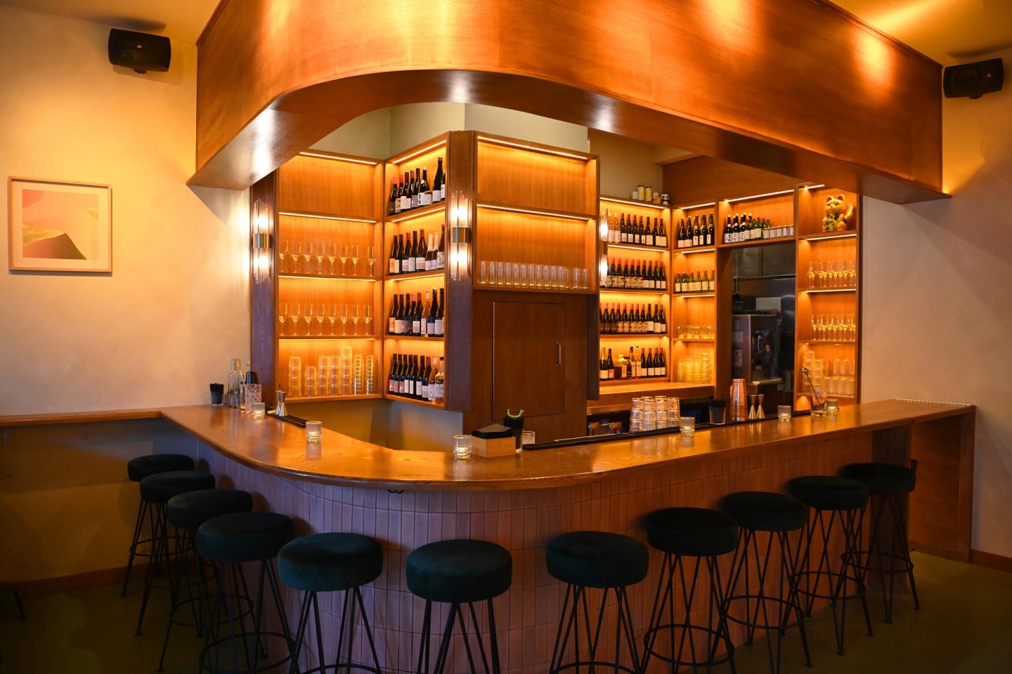 A cozy bar with green stools, curved wooden bar counter, and warm lighting. Shelves behind the bar display wine bottles and glasses. A framed artwork hangs on the left wall.