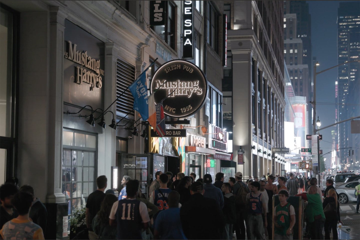A busy nighttime street scene in front of Mustang Harry’s Irish pub in New York City, with a crowd of people walking on the sidewalk and illuminated signs lighting up the area.