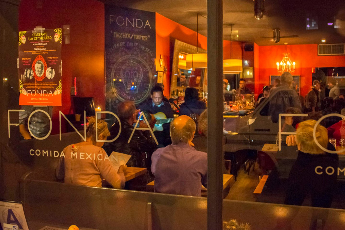 View through the window of a bustling Mexican restaurant, Fonda. Inside, diners are seated at tables enjoying their meals. A musician plays a string instrument in the corner. Warm lighting and decorations create a cozy atmosphere.