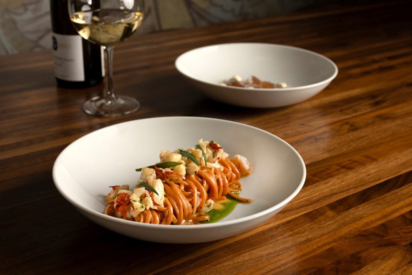 A dish of spaghetti with herbs and seafood is served in a white bowl on a wooden table. Another bowl is in the background. A glass of white wine and a wine bottle are beside the main dish.