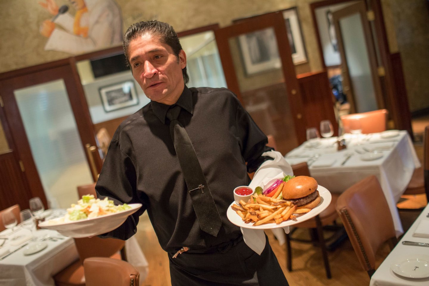 A waiter wearing a black uniform holds two plates in a restaurant. One plate has a burger and fries with ketchup, and the other has a salad. The dining area has white tablecloths, wooden chairs, and framed pictures on the walls.