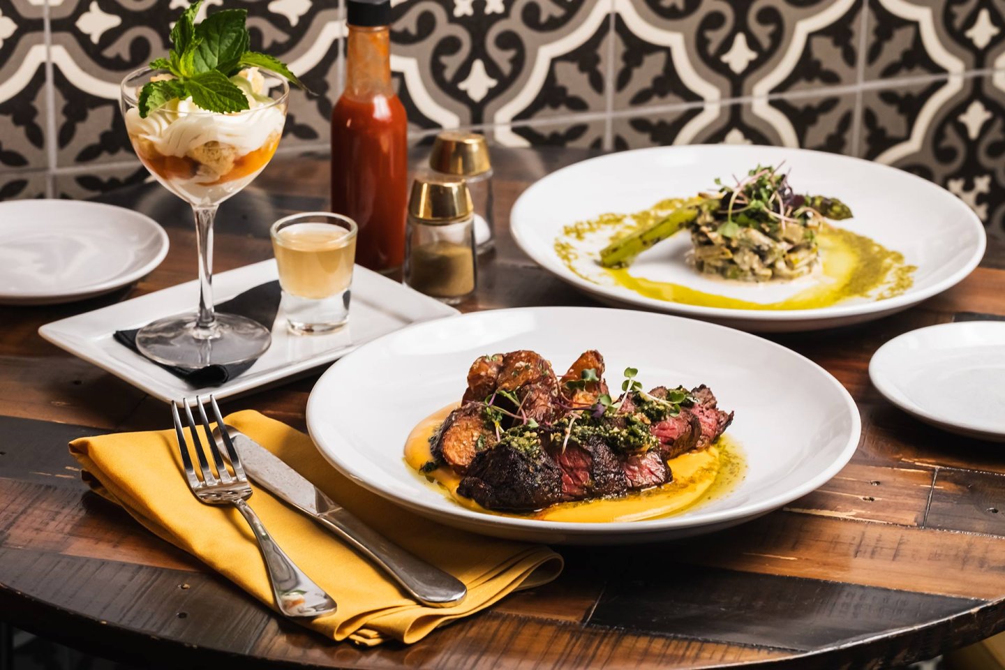 A table set with a plated steak dish, a creamy risotto with asparagus, a dessert topped with mint, a shot glass, and a bottle of hot sauce, against a patterned tile background.