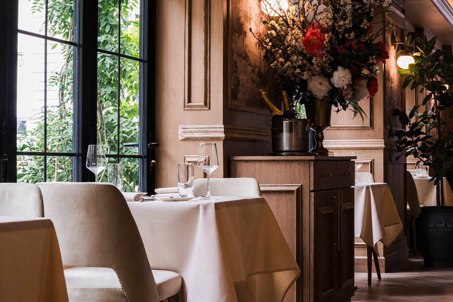 Elegant restaurant interior with cream-colored chairs, tables set with white tablecloths and glassware, large windows letting in natural light, and a floral arrangement on a wooden sideboard.
