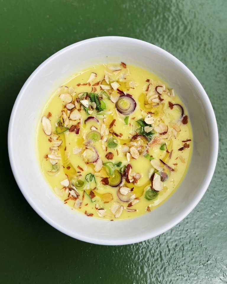 A white bowl filled with yellow soup, garnished with chopped almonds, sliced green onions, and herbs, sits on a textured green surface.