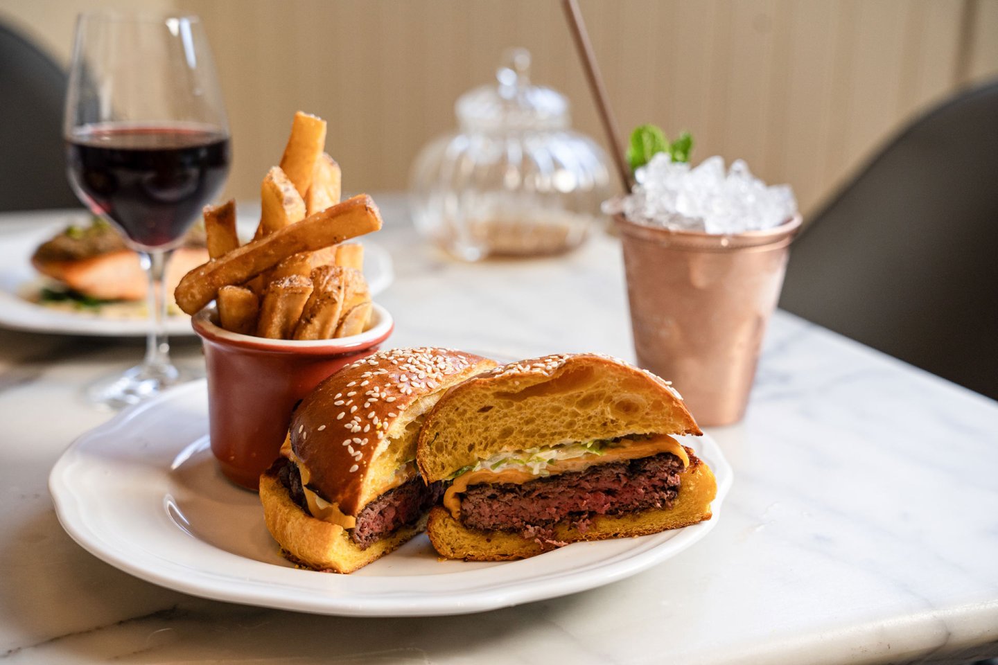 A plate with a sesame seed bun burger cut in half, a cup of French fries, a glass of red wine, and a copper cup with crushed ice and mint on a marble table.