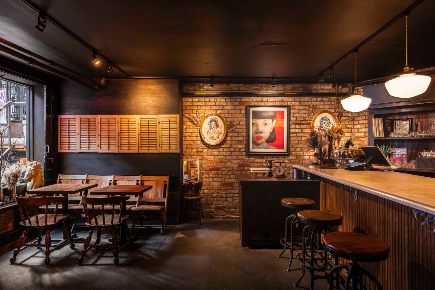 Cozy bar with brick walls, featuring a wooden bar counter and high stools. There are two small tables with chairs by the window. The dimly lit space is decorated with framed art and vintage-style lamps, creating a warm and inviting atmosphere.