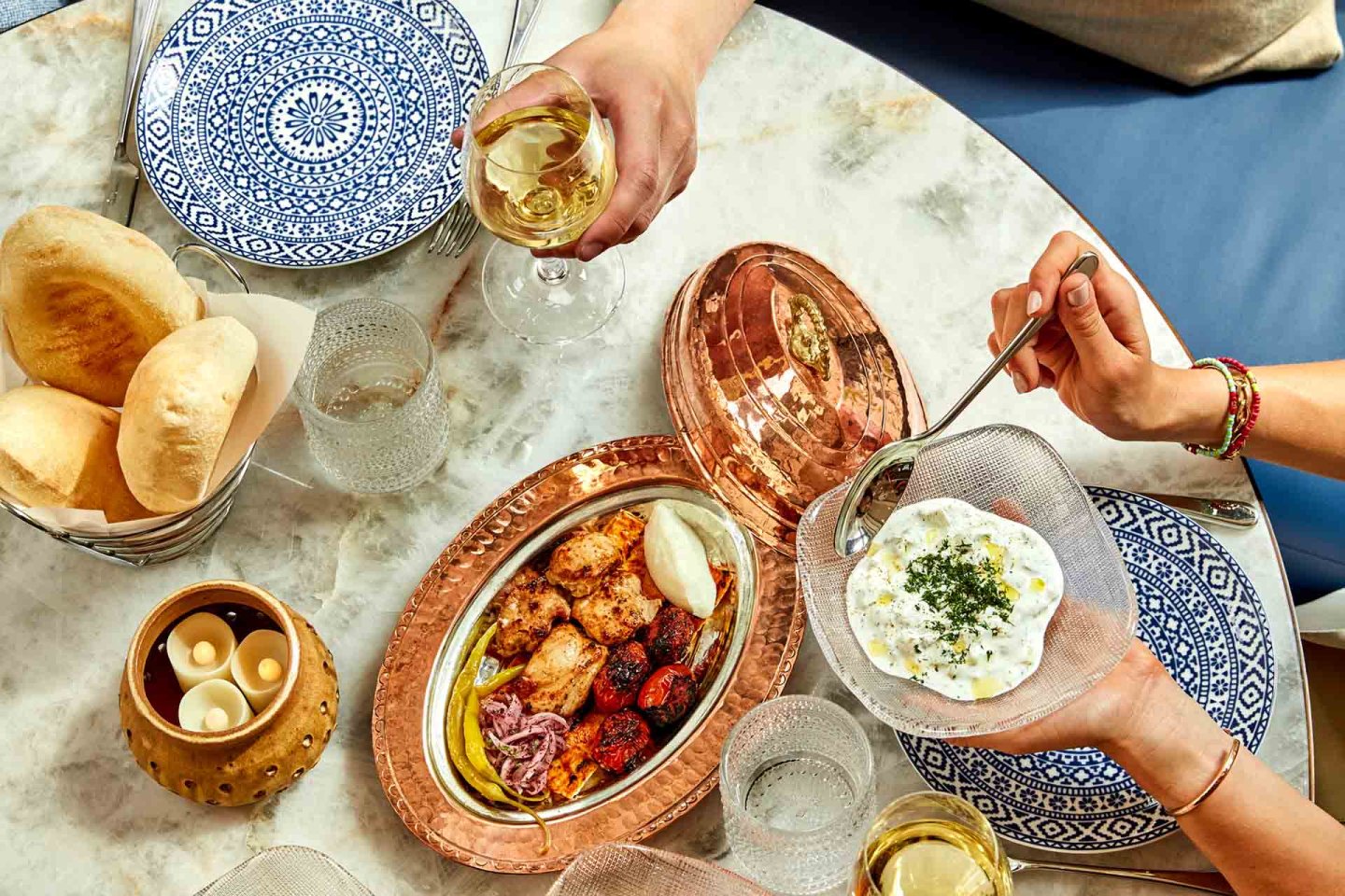 A table set with patterned plates, pita bread, candles, glasses of white wine, and a copper tray holding grilled meats, vegetables, and a bowl of creamy dip being served by hand.