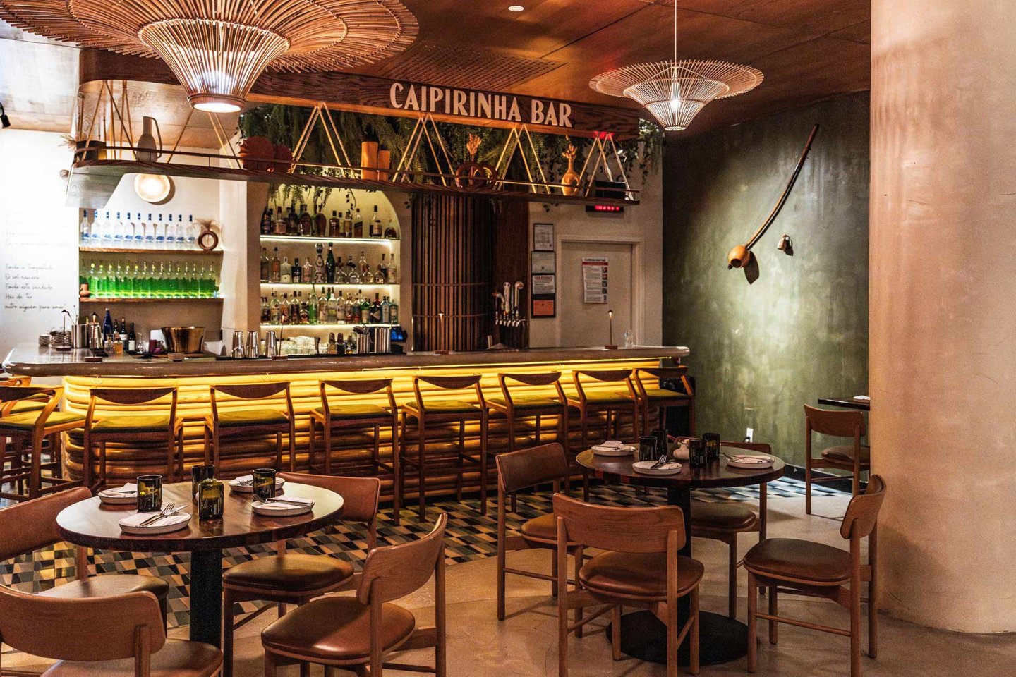 A stylish bar labeled “Caipirinha Bar” with warm lighting, wooden furniture, round tables set for dining, and a well-stocked back bar, creating a cozy and inviting atmosphere.