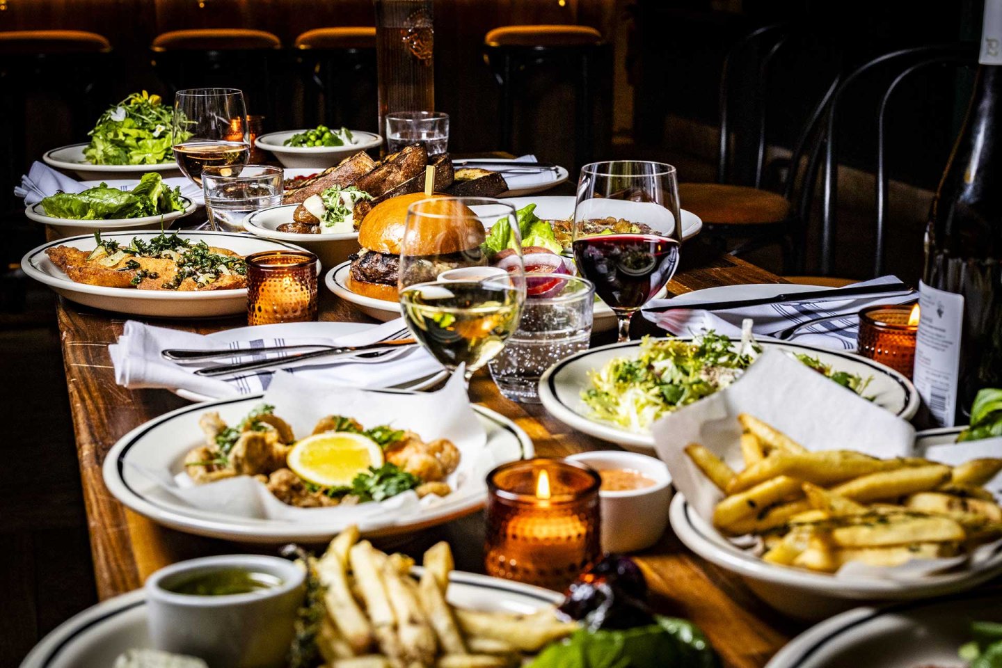 A wooden table set with plates of assorted food including fries, salads, burgers, fish, and grilled meat, accompanied by glasses of wine and water, with candles providing warm lighting.