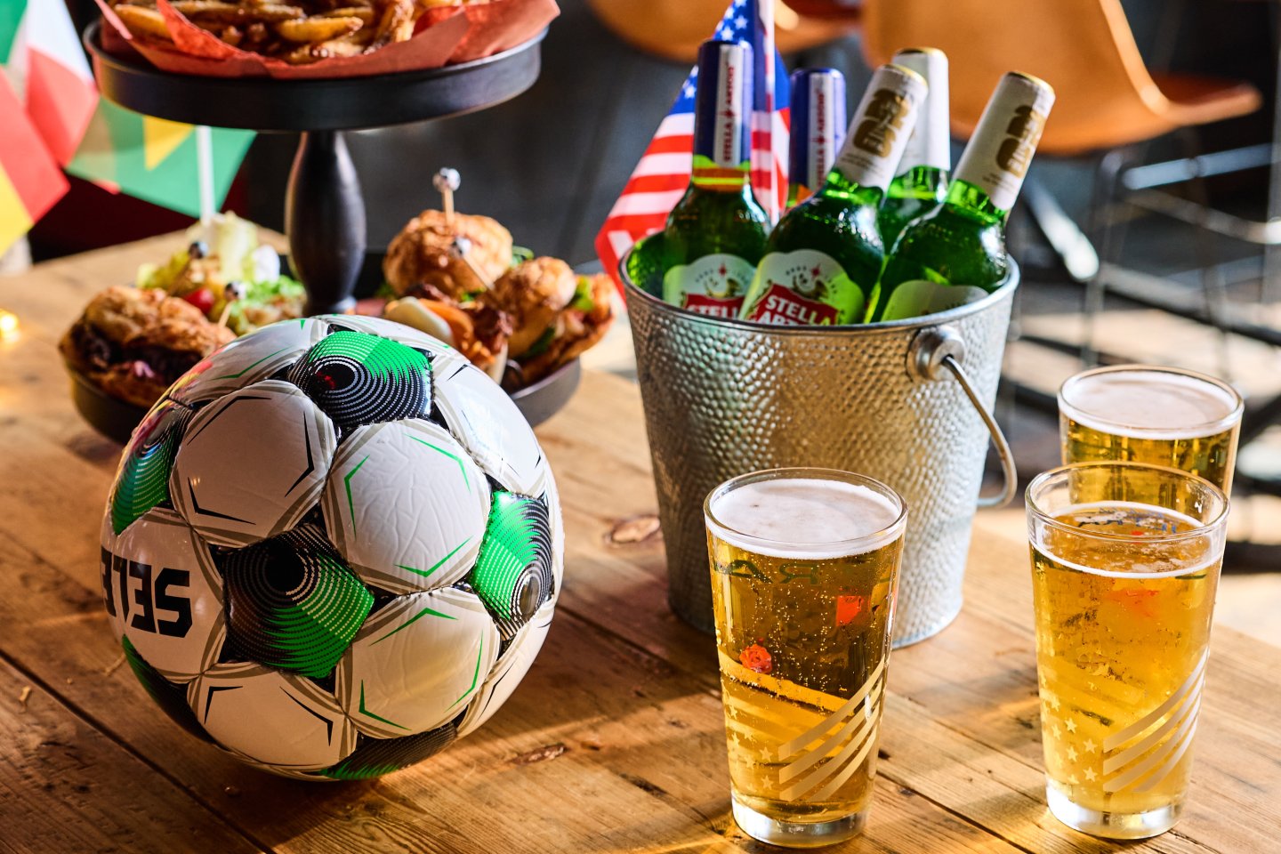 Pitch-Side Pail at BLACKBARN NoMad: six ice-cold Stella Artois bottles in a metal bucket with two golden poured beers, a soccer ball, and an American flag on a wood table, set for a World Cup watch party.