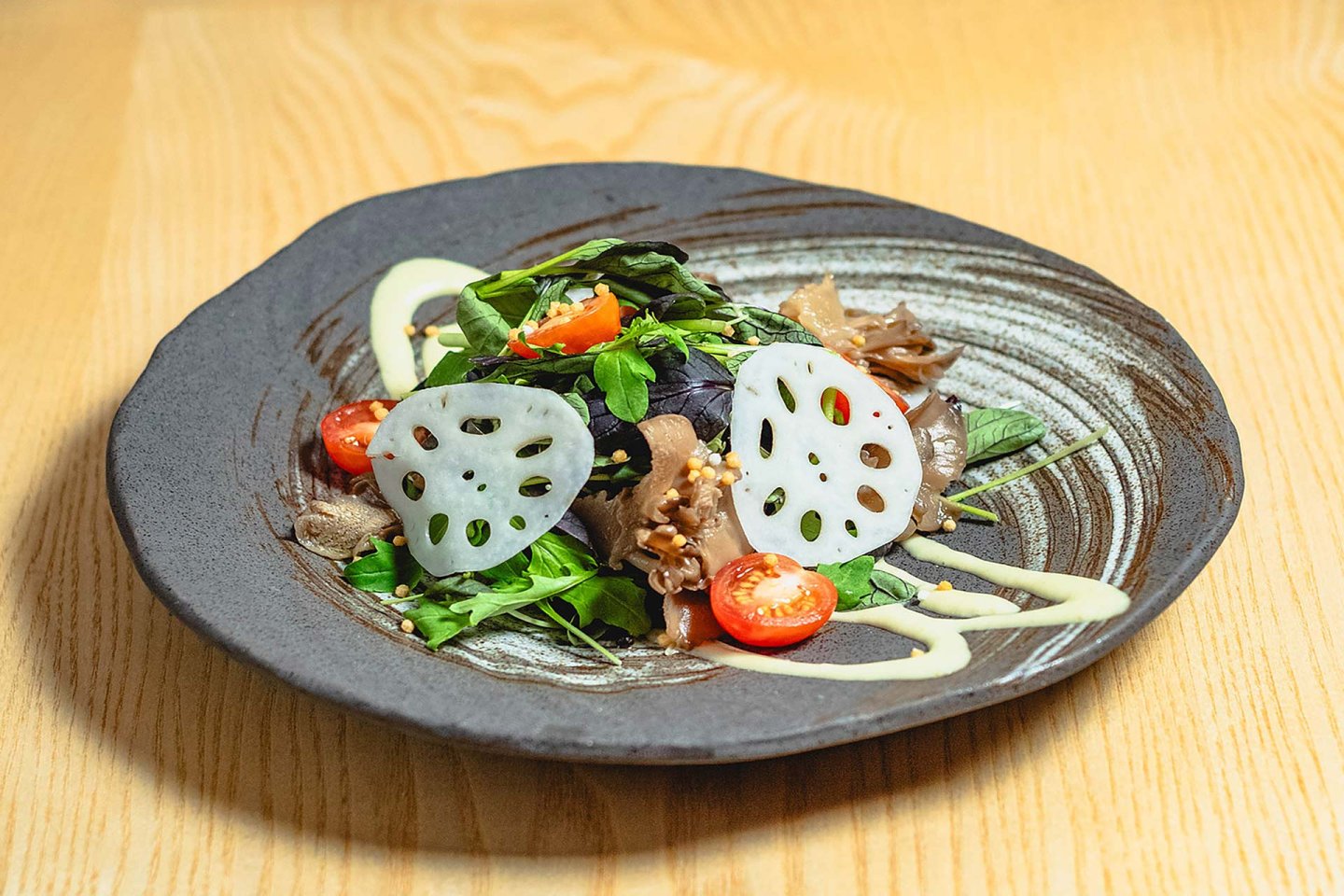 A black ceramic plate with a salad of mixed greens, cherry tomato halves, mushrooms, and thinly sliced lotus root, drizzled with a light dressing, on a light wooden table.