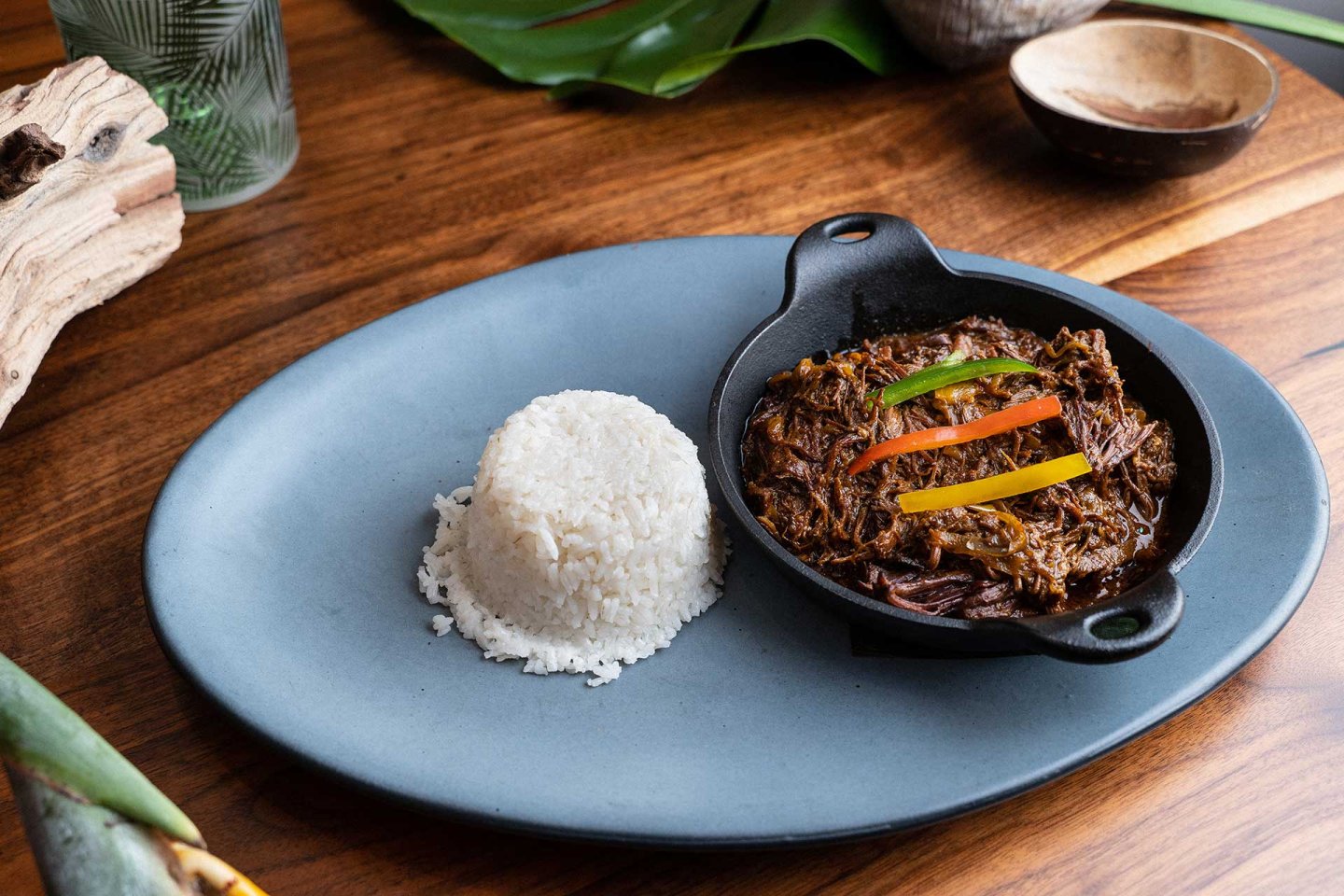 A mound of white rice and a small skillet of shredded beef in sauce, garnished with sliced peppers, are served on a blue oval plate atop a wooden table.
