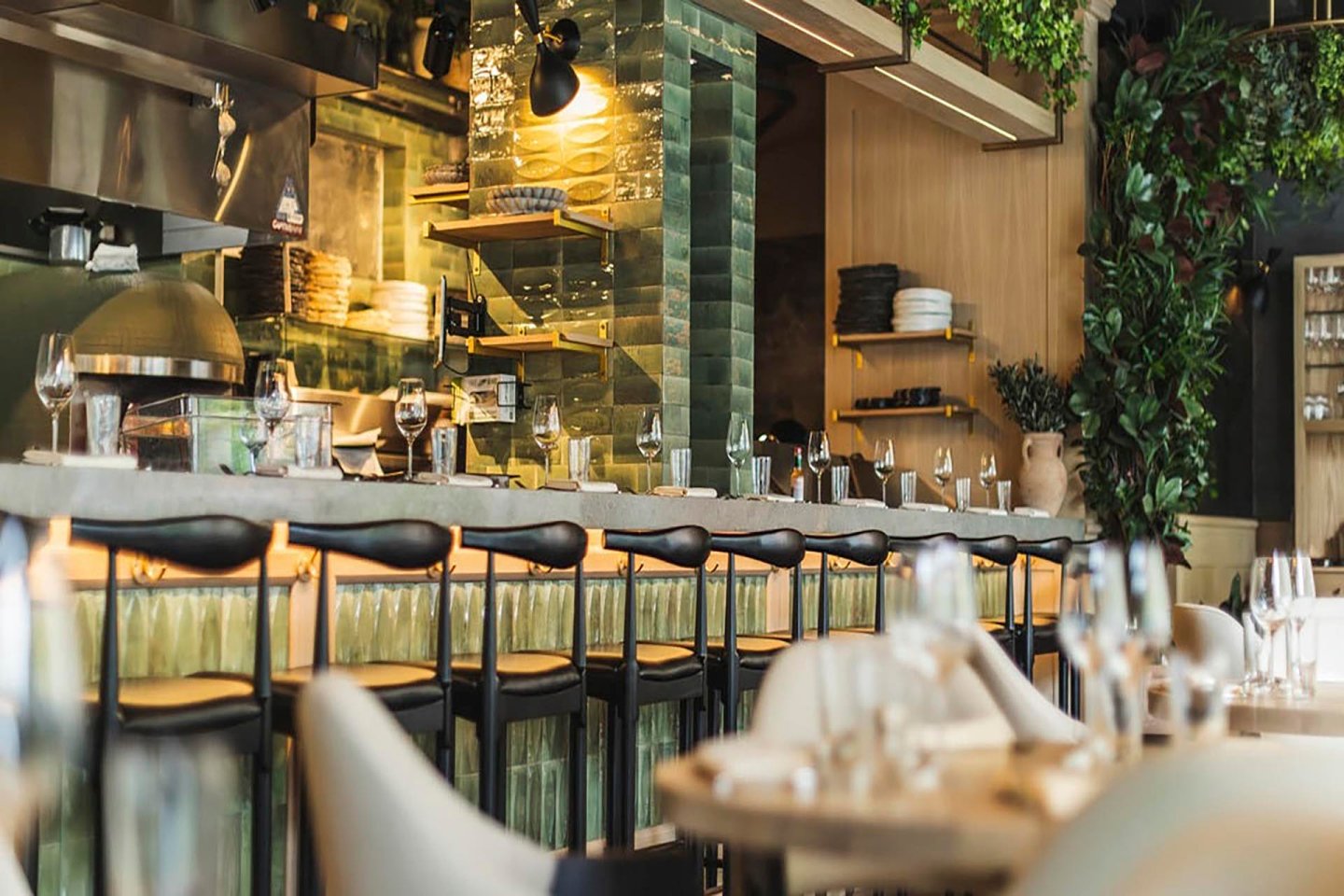 Interior of a modern restaurant featuring a stylish bar with high chairs. Ambient lighting highlights glass bottles and plates on shelves. Tables are set with wine glasses, surrounded by lush greenery on the walls.