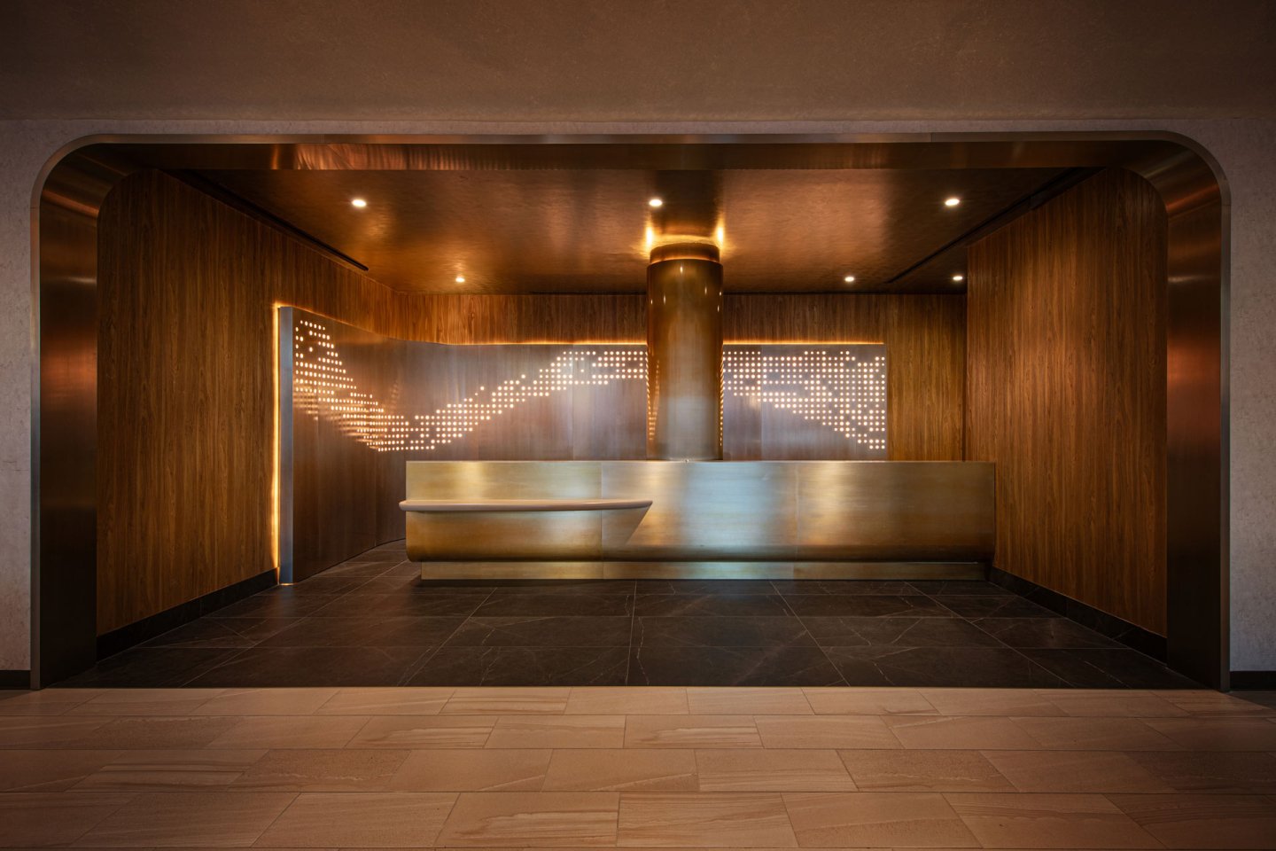 A modern interior with a sleek, wood-paneled reception area and a curved metallic desk. The walls feature artistic backlit designs, creating a warm and elegant ambiance. The floor blends dark and light tiles for a contrasting effect.