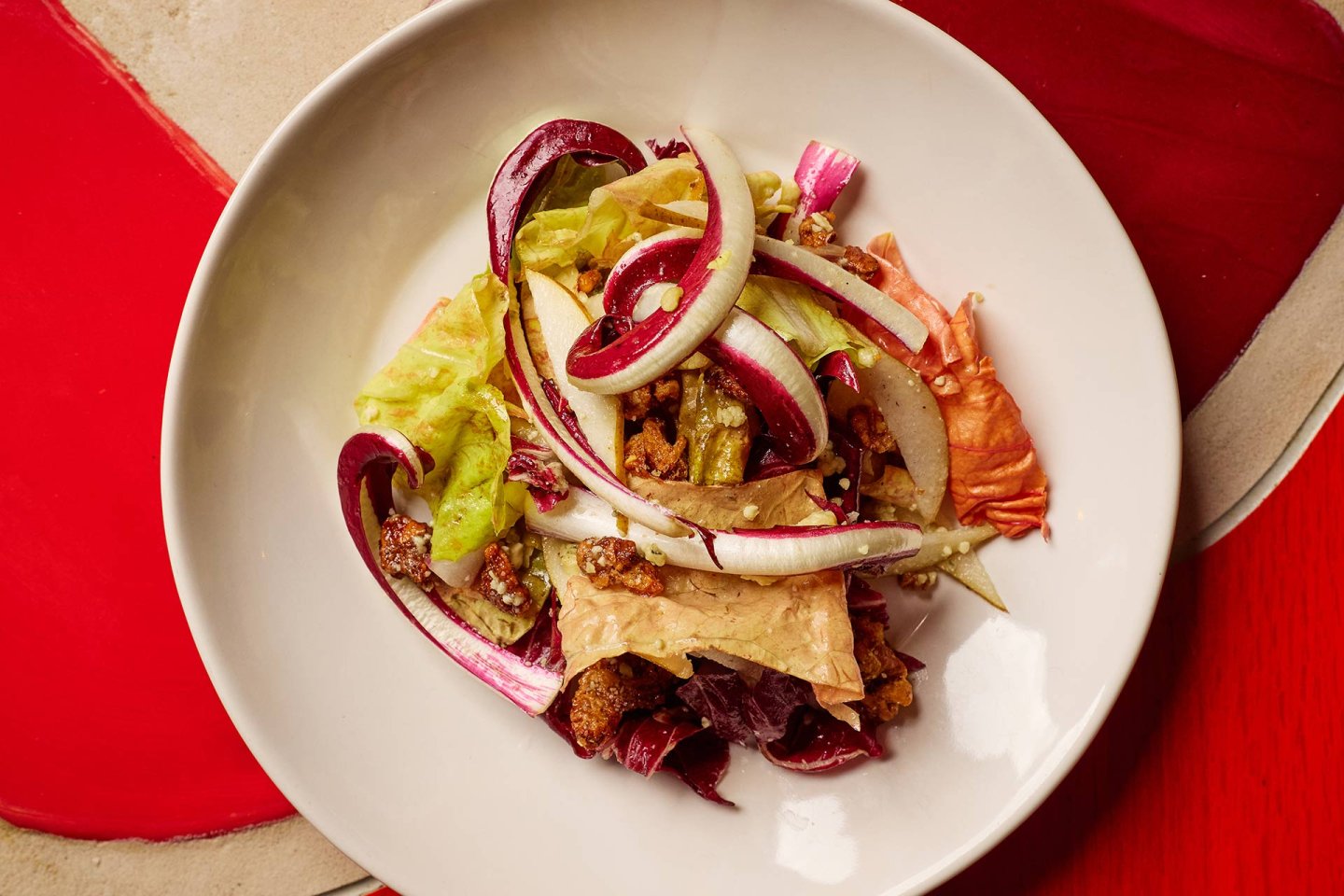 A white bowl filled with a mixed salad of leafy greens, radicchio, sliced onions, crispy croutons, and what appears to be walnuts, set on a table with red and beige placemats.