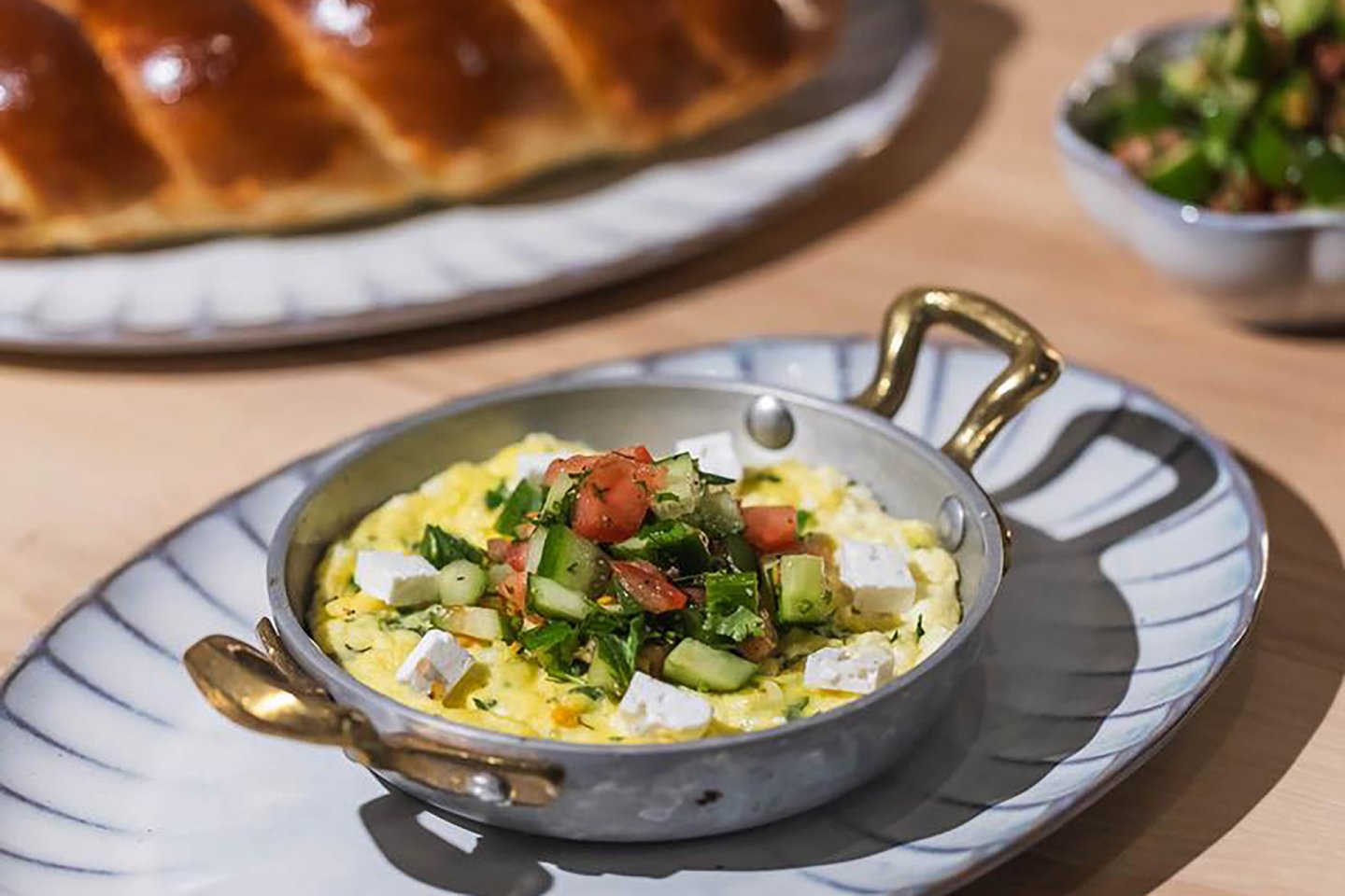 A small brass-handled pan with scrambled eggs topped with diced cucumber, tomato, herbs, and feta cheese sits on a white plate; challah bread and salad are in the background.