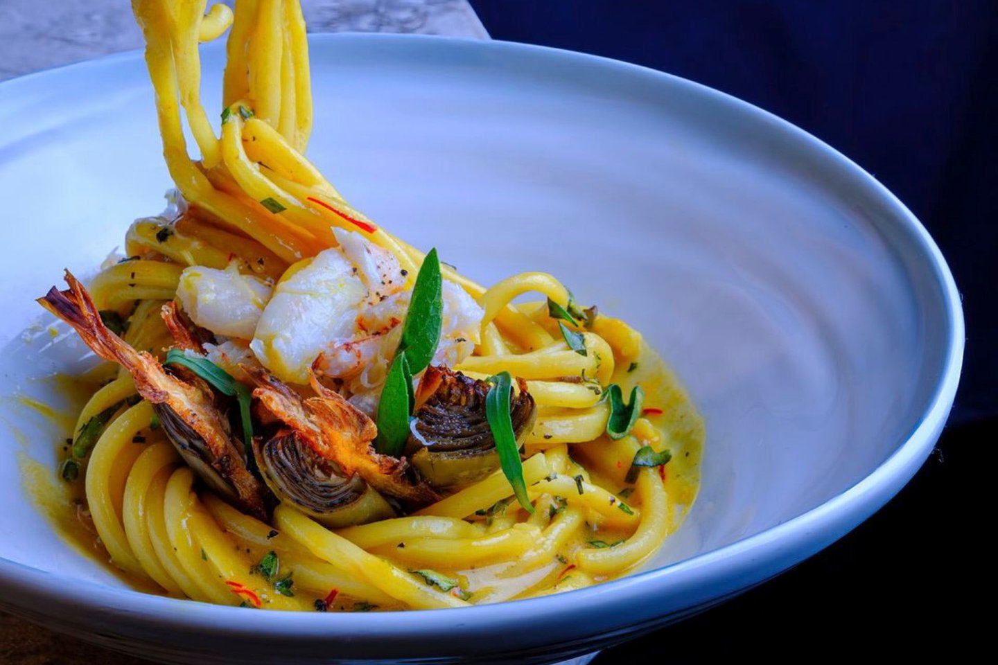 A white bowl filled with bright yellow pasta, garnished with pieces of shrimp, artichoke, and green herbs. The pasta is elegantly twirled. The background is dark, highlighting the vivid colors of the dish.