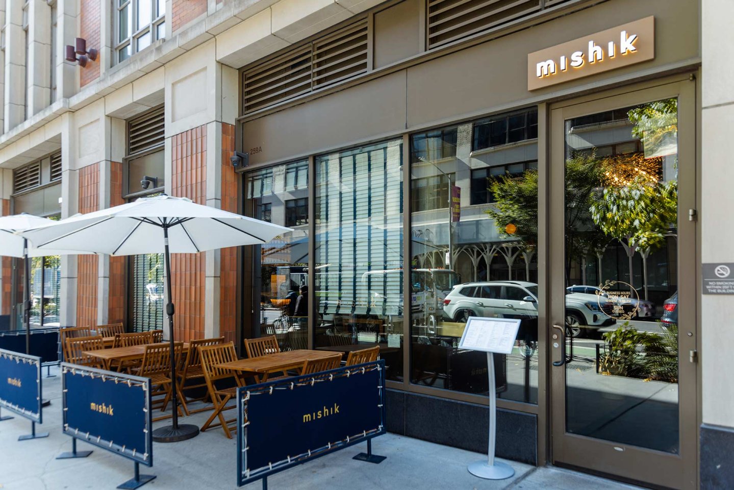 Street view of a restaurant called Mishik with large glass windows, wooden outdoor tables and chairs under white umbrellas, and a menu stand by the entrance. Blue barriers separate the patio from the sidewalk.