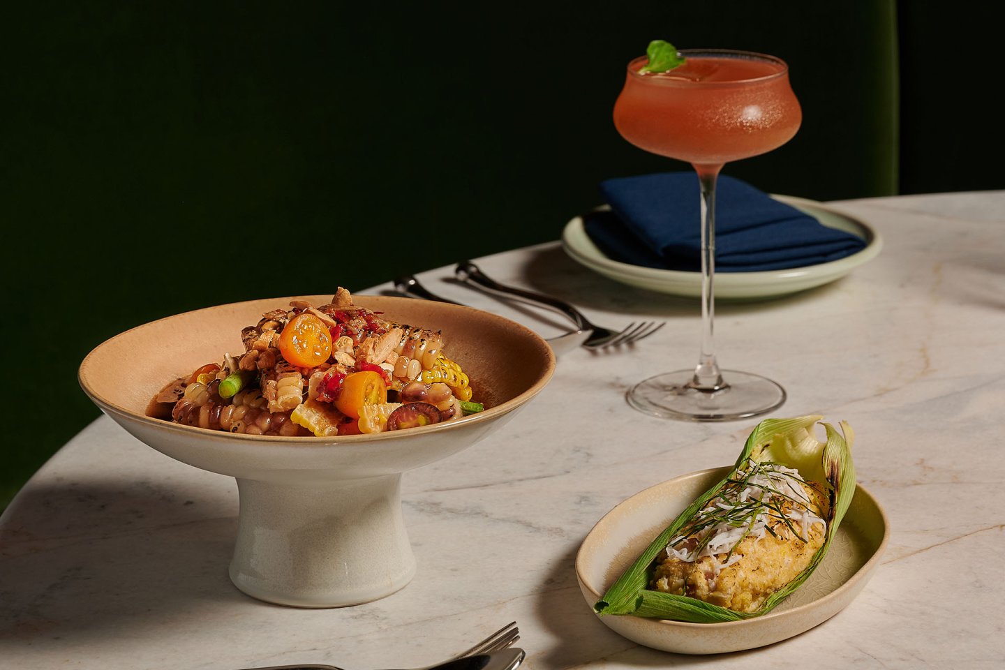 A bowl of colorful vegetable pasta, a stuffed corn husk dish, and a pink cocktail with a mint garnish sit on a marble table with cutlery and a napkin on a plate in the background.