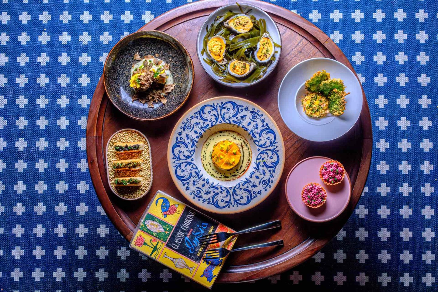 A round wooden table with six colorful plates of assorted gourmet appetizers, two spoons, and a vibrant illustrated menu, on a blue patterned tablecloth.