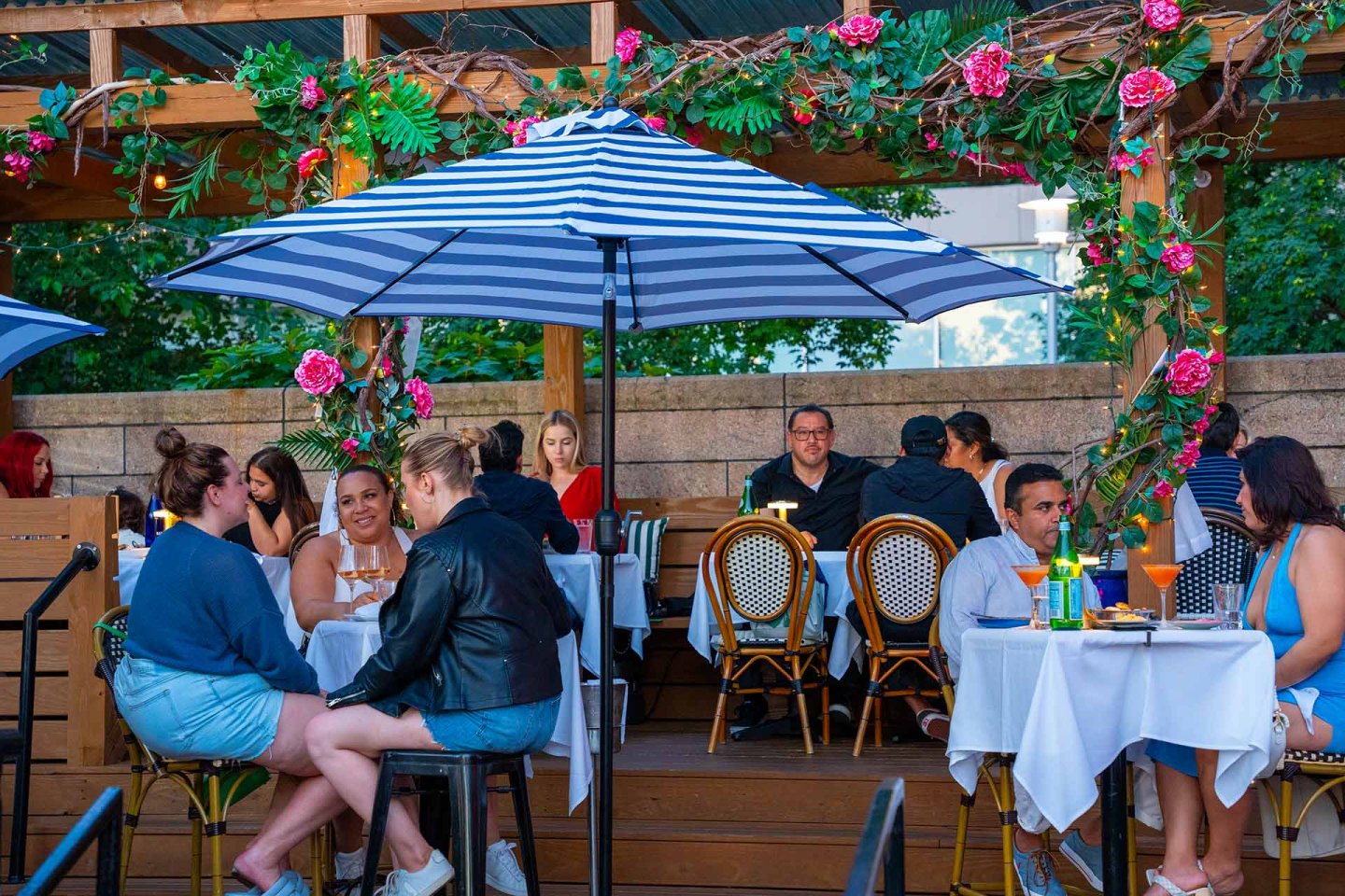People dining outdoors at a restaurant patio with striped umbrellas, white tablecloths, and floral decorations. Groups are seated at tables, talking and enjoying food and drinks in a lively, colorful setting.