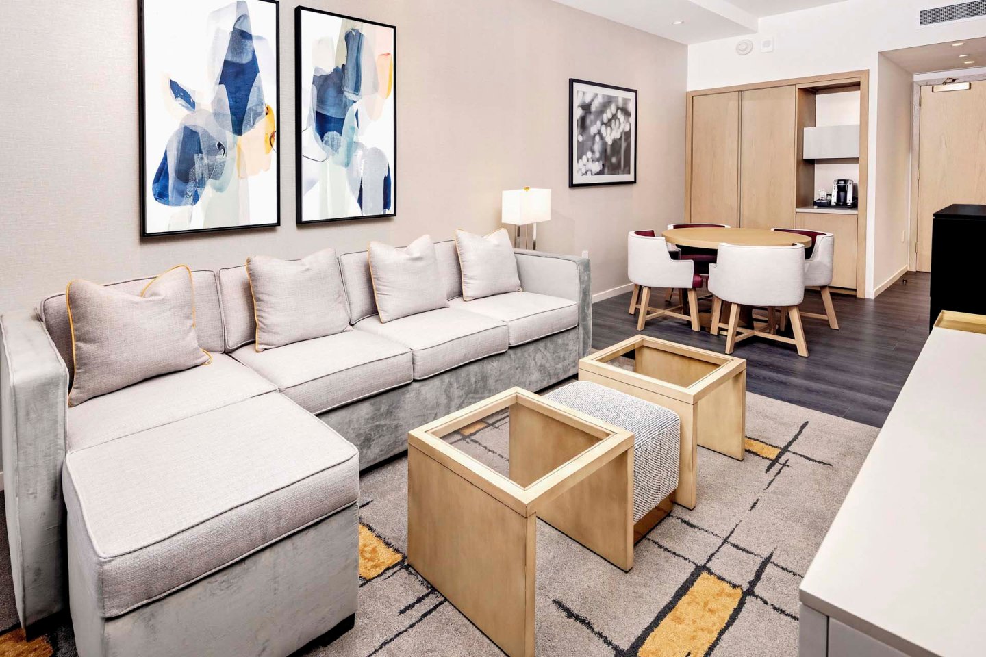 A modern living room with a large gray sectional sofa, two wooden coffee tables, and abstract art on the wall. A small dining area with a round table and four chairs is in the corner, next to a kitchenette. The room has a contemporary decor.
