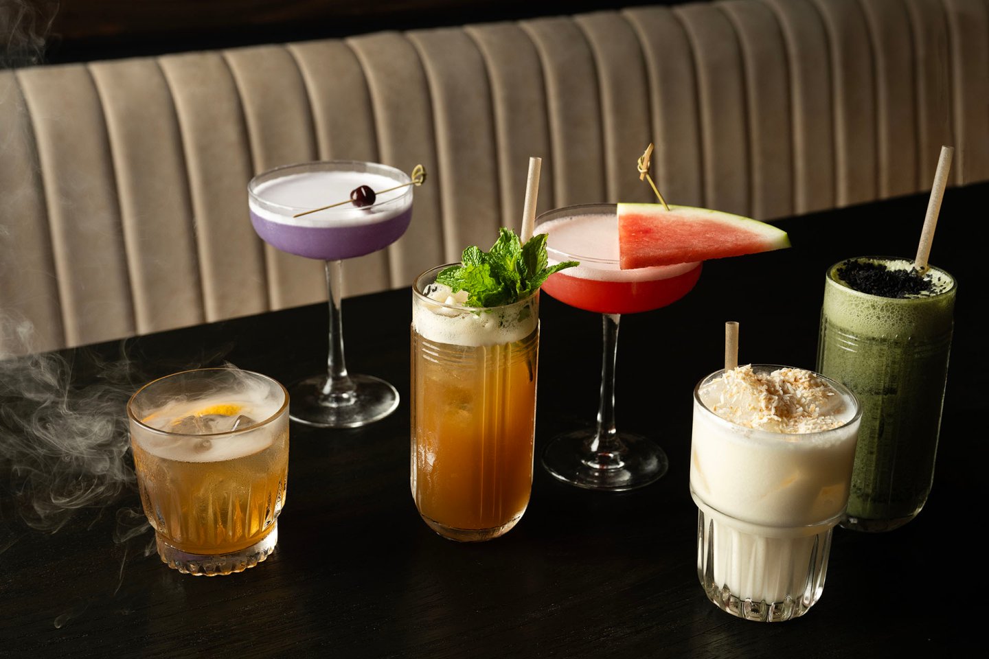Six assorted cocktails on a dark table, featuring vibrant colors and garnishes like mint, a cherry, a watermelon slice, a smoky effect, and whipped cream; booth seating in the background.