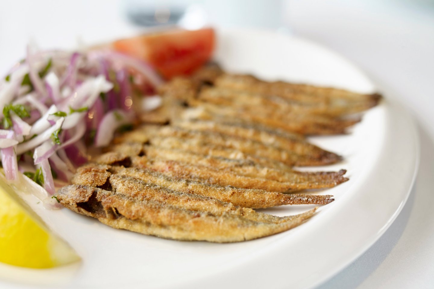 A plate of fried small fish served with a lemon wedge, sliced red onions, herbs, and a tomato slice on the side.