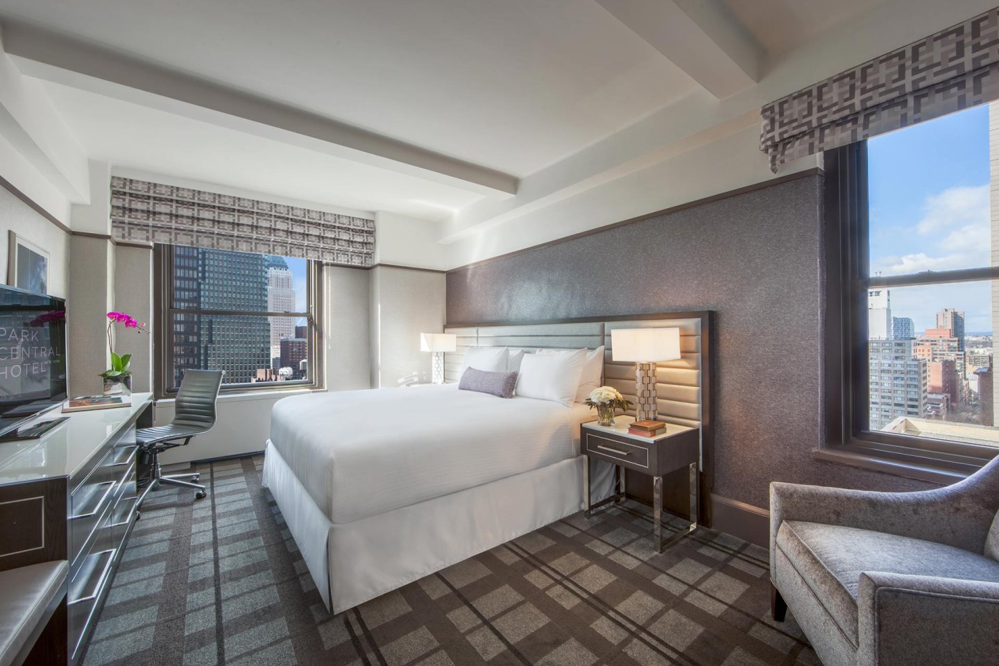 Modern hotel room with a large bed, two nightstands, a desk with a chair, a television, and a gray armchair. Large windows reveal a cityscape with tall buildings and a blue sky outside.