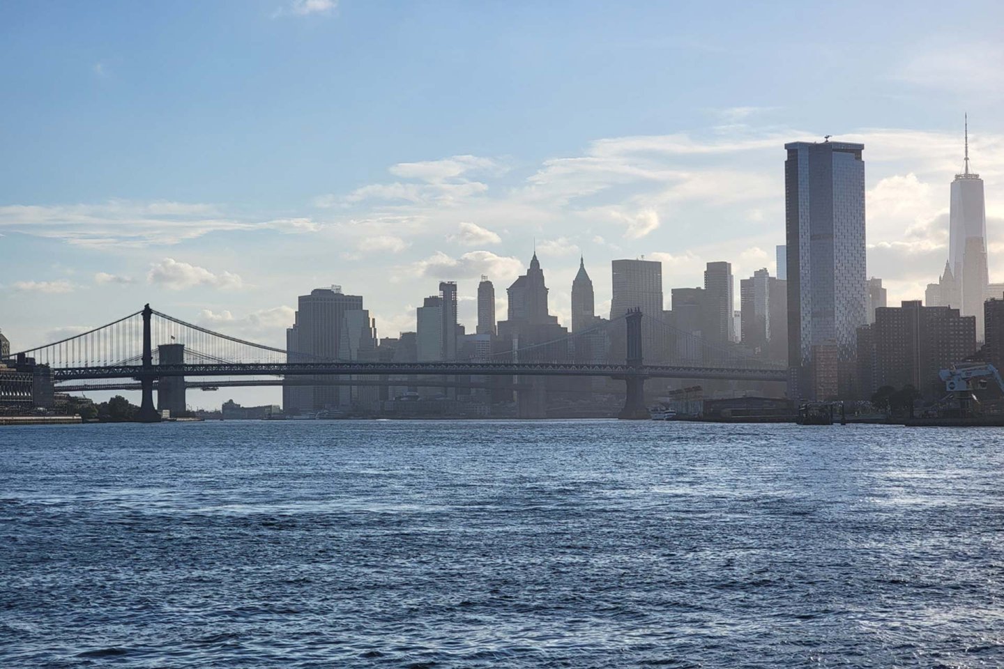 A scenic view of a city skyline at dusk, showcasing tall skyscrapers including a prominent tower and a suspension bridge over a river. The sky is partly cloudy with the setting sun casting a soft light on the buildings.