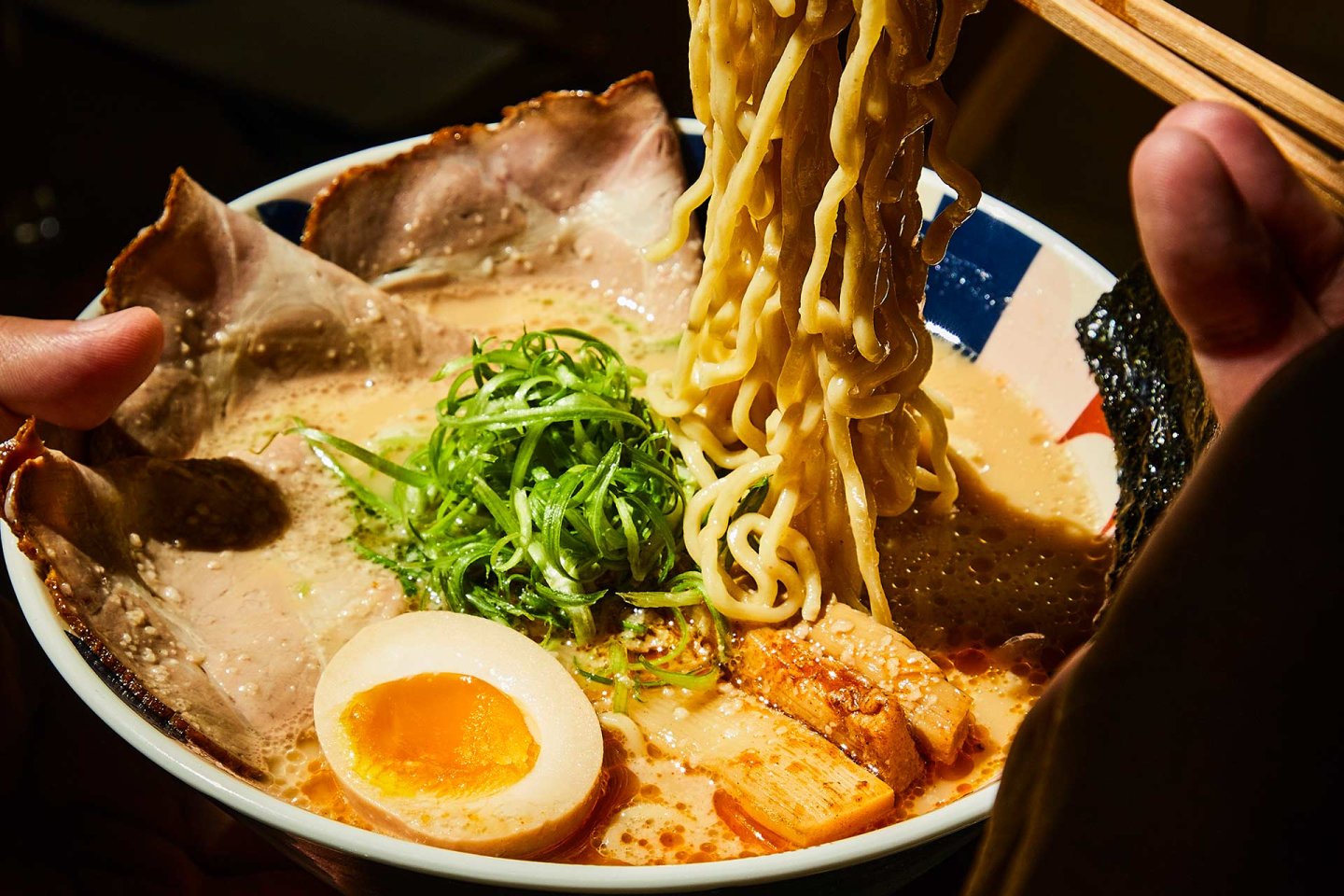 A bowl of ramen with thick noodles being lifted by chopsticks, slices of pork, half a soft-boiled egg, green onions, bamboo shoots, seaweed, and rich broth.
