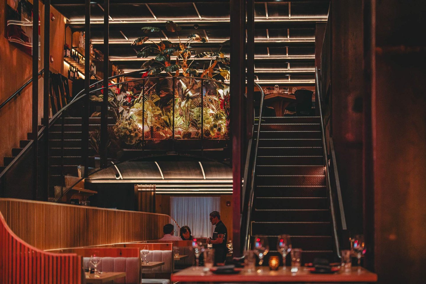 Dimly lit restaurant interior with modern decor, featuring an elevated area with lush plants. A few diners are seated at tables, and a waiter is visible. Ambient lighting and stylish design create a cozy and elegant atmosphere.