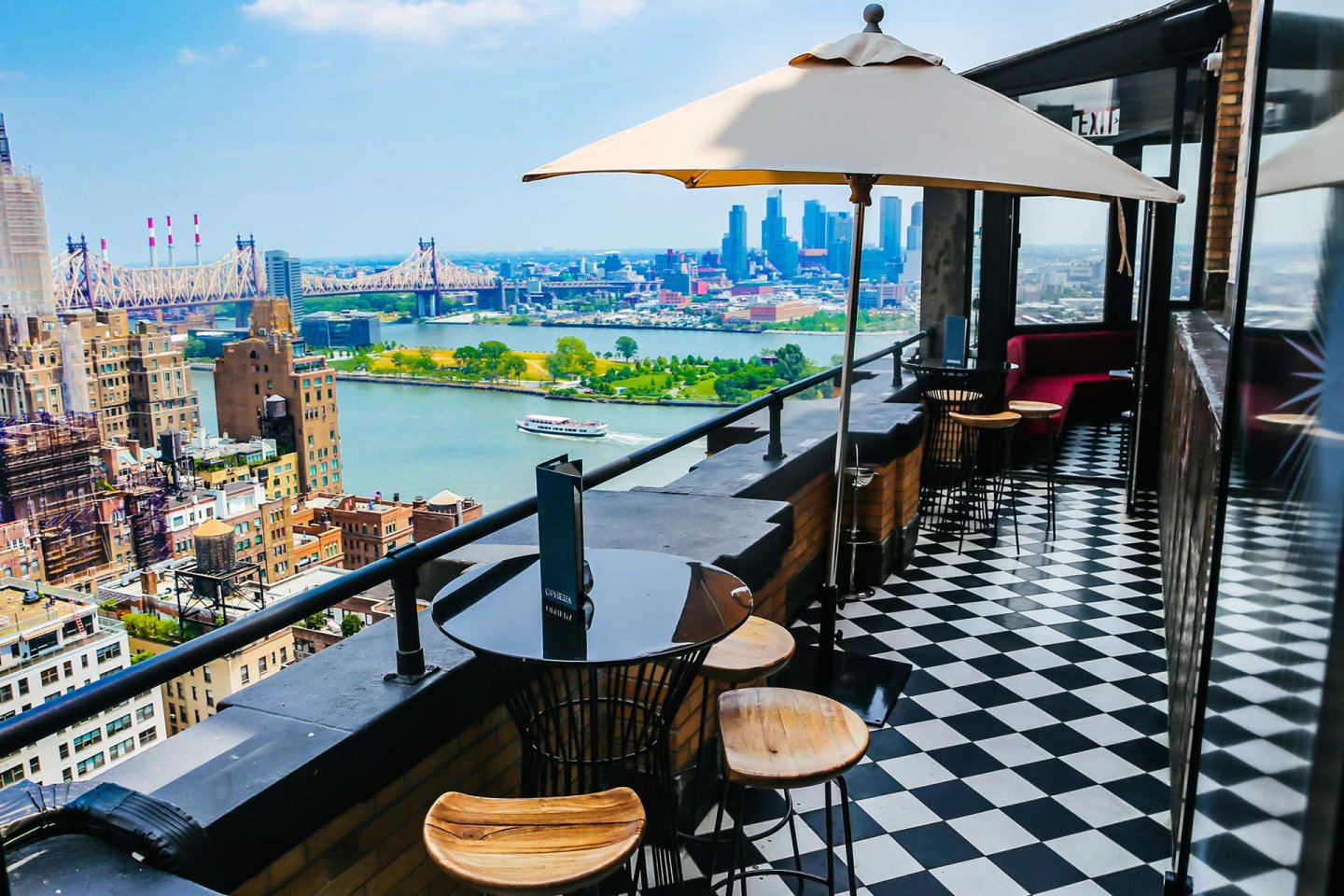 Rooftop bar with black and white checkered floor, round tables, wooden stools, and a large umbrella overlooking a cityscape, river, bridge, and distant skyline on a sunny day.