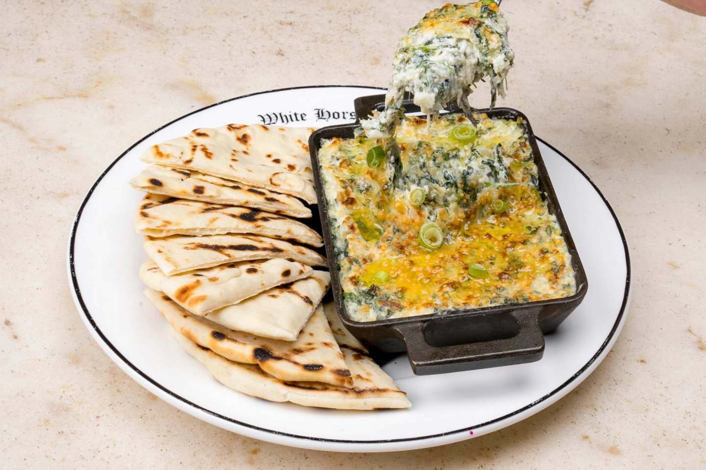 A plate with grilled flatbread slices on the left and a small skillet of spinach and cheese dip topped with green onions on the right. A hand holds a scoop of the dip above the skillet. The plate has "White Horse" written on the rim.