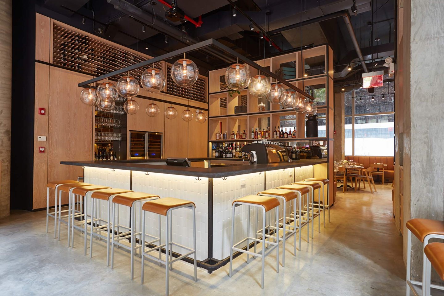 Modern bar with light wood paneling, globe pendant lights, white tiled bar counter, and tan leather stools. Shelves hold bottles and glassware; tables are visible in the background. Large windows let in natural light.