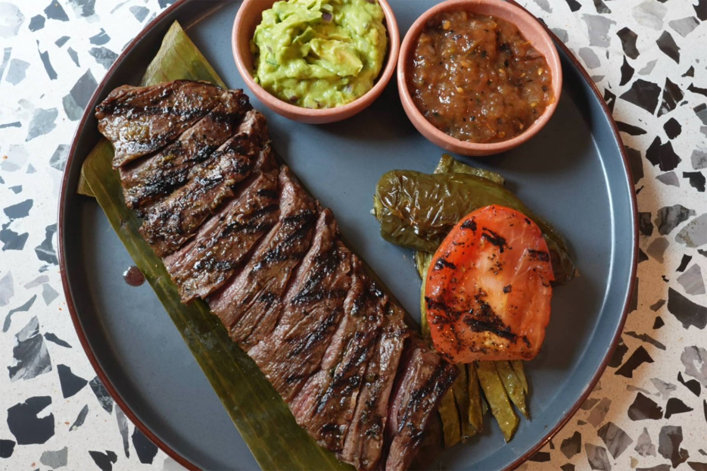 A plate with grilled steak on a banana leaf, served with guacamole, salsa, a grilled jalapeño, grilled tomato, and cactus on a terrazzo surface.