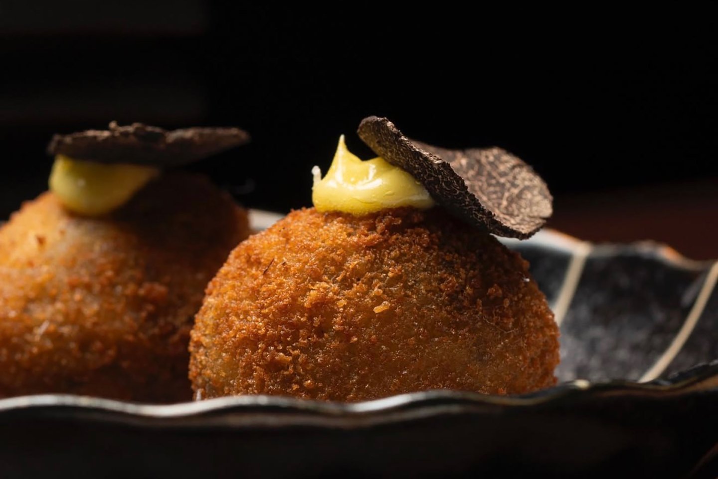 Two golden-brown, crispy croquettes topped with a dollop of yellow sauce and a thin slice of black truffle, served on a dark, decorative plate.