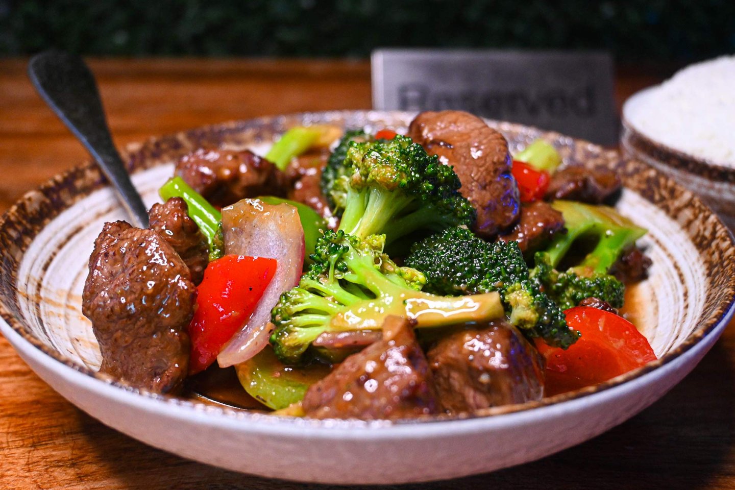 A bowl of stir-fried beef with broccoli, red bell peppers, and onions in a savory sauce, served with a spoon. There is a small bowl of white rice and a "Reserved" sign in the background.