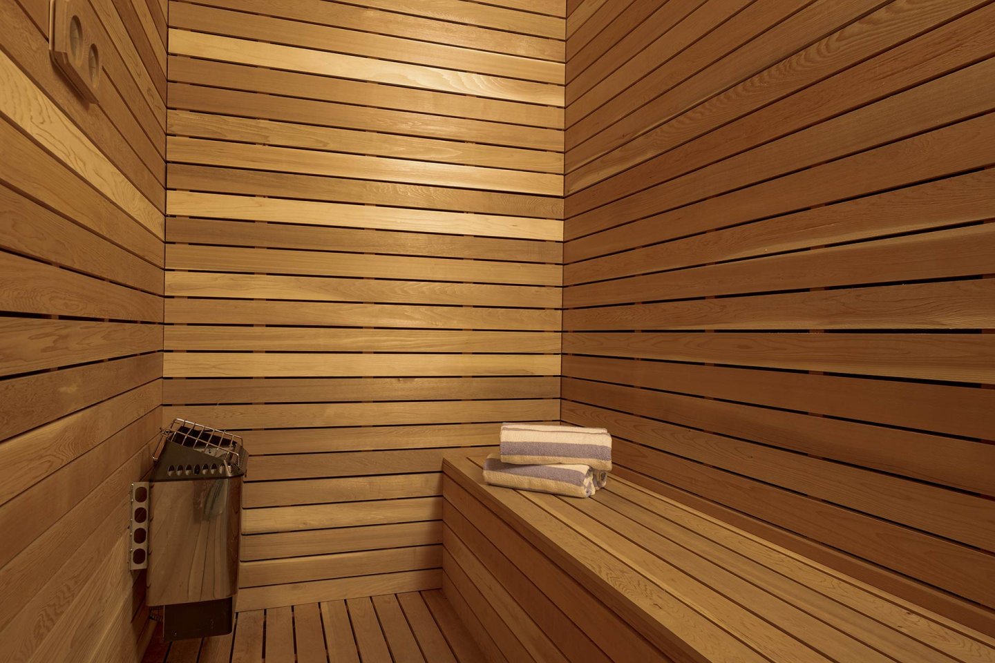 A wooden sauna interior with light-colored horizontal wood panels, a bench, neatly folded towels, and a metal heater in the corner. The space appears clean and inviting.
