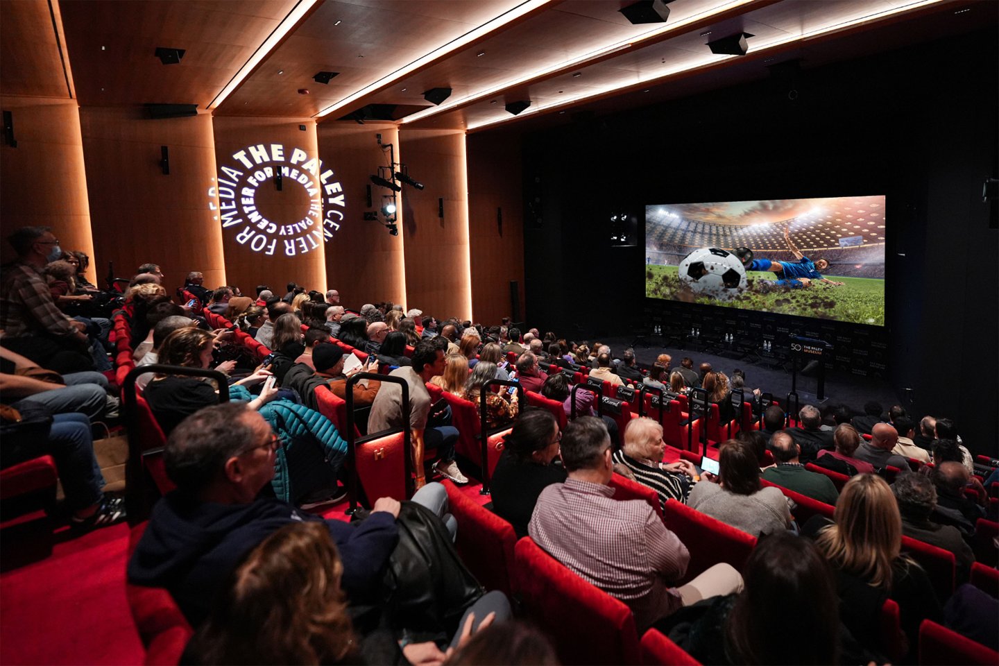Audience seated in a darkened theater at the Paley Museum, watching a large screen showing a soccer player sliding on the field next to the ball